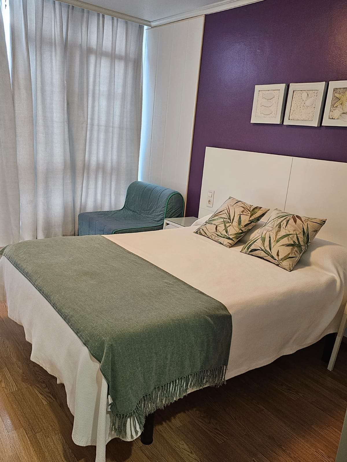 habitación doble con una cama de matrimonio con mantas de color verde y blanco, y la pared en color violeta. La cama está hecha y tiene varios cuadros de decoración encima de la misma