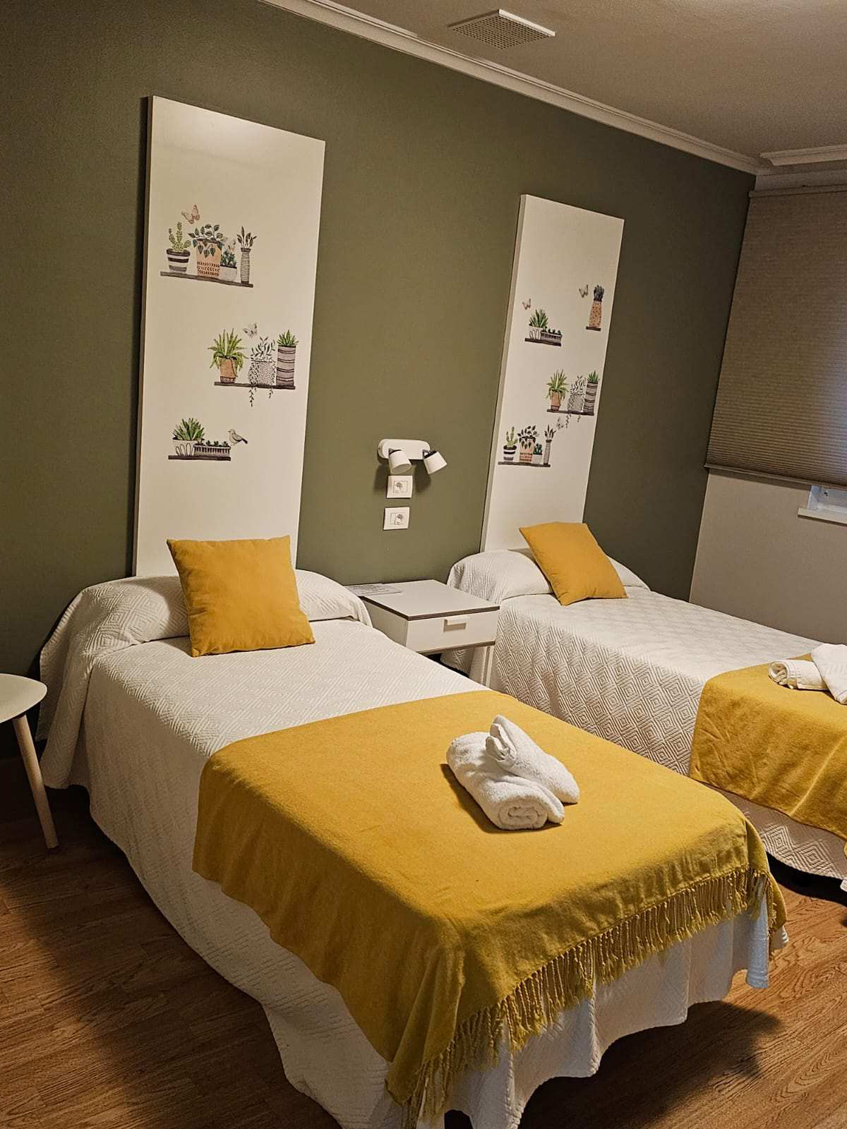 habitación doble con dos camas con mantas de color amarillo y la pared en color verde. Hay dos camas hechas con toallas en forma de muñecos encima de la misma. encima de la cama hay decoración en tonos blancos.